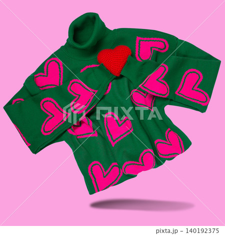 Green knitted turtleneck sweater with pink hearts pattern hugging a small red handmade heart, levitating on pink background. Green knitted turtleneck sweater with pink hearts pattern hugging a small red handmade heart, levitating on pink background. 140192375