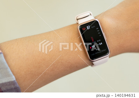 Smartwatch displays heart rate and activity data. Wristband is light pink, sleek and modern. User wears casual clothing, relaxed posture. Screen shows vibrant red and black digital interface 140194631