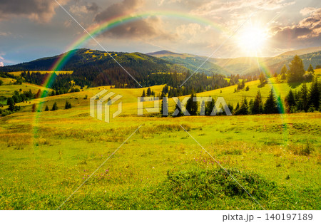 sunset over mountainous countryside landscape in summer. view of green rural scenery with grassy pastures and rolling forested hills. wonderful mountain weather with cloudy sky. weather with rainbow 140197189