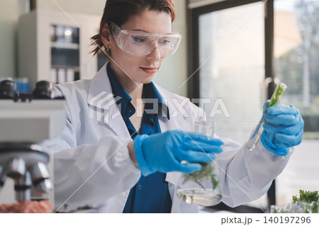 Scientists in lab coats work with plants in a modern lab, focusing on research and experimentation under bright, sterile conditions. 140197296
