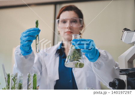 Scientists in lab coats work with plants in a modern lab, focusing on research and experimentation under bright, sterile conditions. 140197297