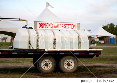A white mobile drinking water station tank trailer parked at an outdoor festival field with white event tents 140197432
