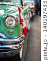 A classic car show is happening in downtown. People are walking around and looking at vintage vehicles. A green convertible is parked with Canadian flags and signs. 140197433
