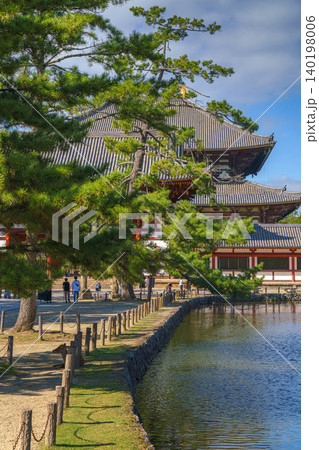Nara, Japan - Sep 26 2024, vertical view across the pond and trees to the facade of Todaiji Temple, Great Buddha Hall, with a blue cloudy sky, with people on background, Nara, Japan Nara, Japan - Sep 26 2024, vertical view across the pond and trees to the facade of Todaiji Temple, Great Buddha Hall, with a blue cloudy sky, with people on background, Nara, Japan 140198006
