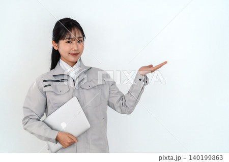 ノートPCを抱え手のひらで案内するグレーの作業着姿の若い女性技術者 白背景 ノートPCを抱え手のひらで案内するグレーの作業着姿の若い女性技術者 白背景 140199863