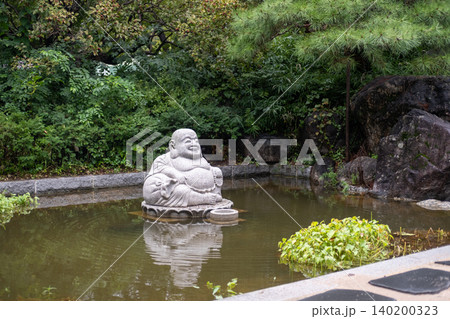 Stone Budai, or Laughing Buddha statue sitting in a shallow pond at Bongeunsa Temple in Seoul, surrounded by calm water and lush greenery. Stone Budai, or Laughing Buddha statue sitting in a shallow pond at Bongeunsa Temple in Seoul, surrounded by calm water and lush greenery. 140200323