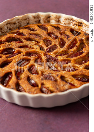 A plum pie is placed on a table with thinly sliced plums visible on top. Bowls of fresh plums are nearby with empty plates waiting for dessert 140201953