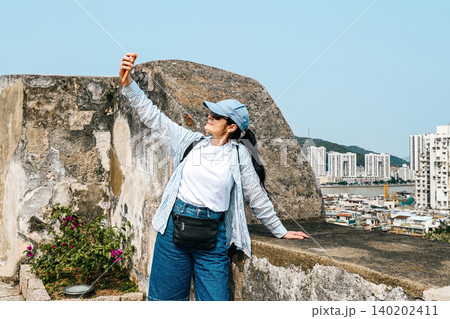 Mature 50 years old Russian woman tourist in cap taking selfie with phone at unesco monte fortress with cityscape panorama view during travel in Macau 140202411
