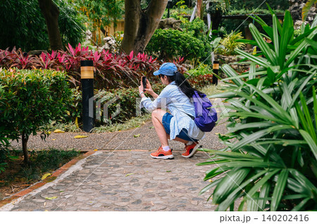 Mature 50 years old Russian woman tourist in cap taking photo with phone at beautiful Lou Lim Ioc Garden during solo travel vacation in Macau China 140202416
