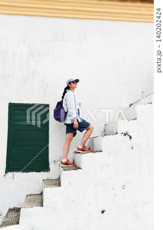 Mature 50 years old Russian woman tourist in cap walking up ancient concrete stairs near green window at Guia Lighthouse area in Macau destination 140202424