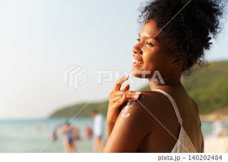 Beautiful African woman applying sunscreen on her shoulder at the beach 140202484