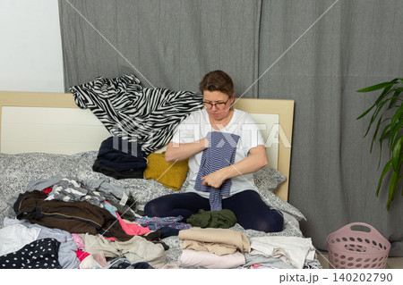 A 52-year-old woman sorts laundry on a bed. She holds a shirt and organizes various garments scattered around. A laundry basket sits nearby as she works on the task at hand A 52-year-old woman sorts laundry on a bed. She holds a shirt and organizes various garments scattered around. A laundry basket sits nearby as she works on the task at hand 140202790