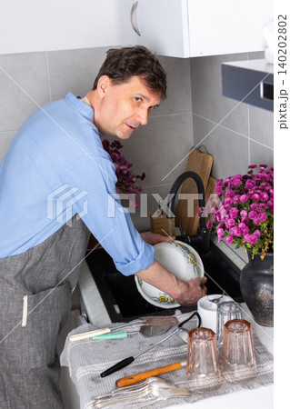 A man stands at a sink in a kitchen, washing dirty dishes and glassware. Flowers sit on the counter as fresh light comes in through the window. The setting is busy and practical A man stands at a sink in a kitchen, washing dirty dishes and glassware. Flowers sit on the counter as fresh light comes in through the window. The setting is busy and practical 140202802