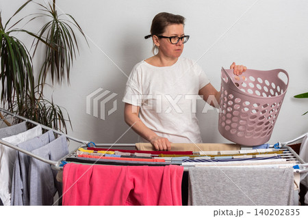 A 50-year-old woman hangs freshly washed laundry on a drying rack inside her room. She is holding a laundry basket filled with clothes, organizing her space 140202835