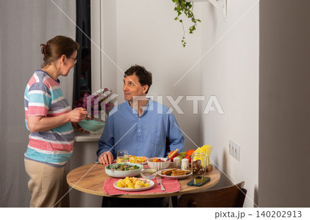A married couple sits at a round table eating dinner. The woman serves food to the man who is smiling. The setting is cozy with a few dishes on the table in the evening A married couple sits at a round table eating dinner. The woman serves food to the man who is smiling. The setting is cozy with a few dishes on the table in the evening 140202913