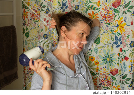 A woman in her fifties stands in front of a colorful floral background. She is drying her hair with a hair dryer. It is morning and she is preparing for the day ahead 140202914