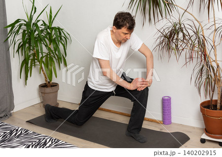 A 53-year-old man performs exercises in his living room at home, stretching and working on his body movements. Green plants are visible in the background adding to the atmosphere A 53-year-old man performs exercises in his living room at home, stretching and working on his body movements. Green plants are visible in the background adding to the atmosphere 140202915