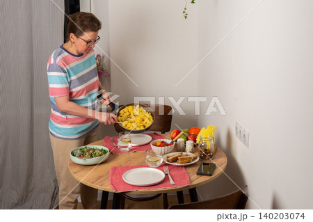 A 53-year-old woman prepares dinner at a round wooden table in her kitchen. She pours food from a colander onto a plate while arranging utensils and other items A 53-year-old woman prepares dinner at a round wooden table in her kitchen. She pours food from a colander onto a plate while arranging utensils and other items 140203074