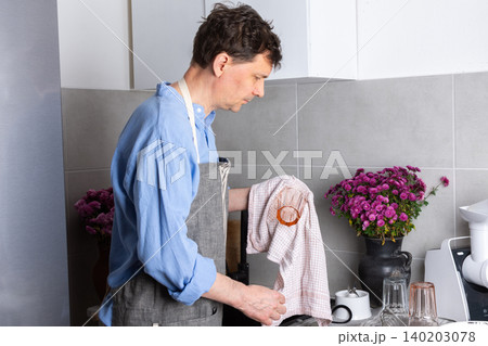 A man stands in his kitchen, drying clean dishes with a cloth. He carefully wipes a plate while surrounded by other washed items on the counter 140203078