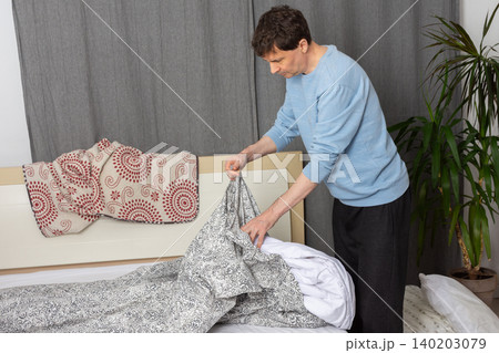 A 53-year-old man is in his bedroom changing bed linen. He holds a patterned bed cover and a white duvet as he prepares to make the bed clean and tidy A 53-year-old man is in his bedroom changing bed linen. He holds a patterned bed cover and a white duvet as he prepares to make the bed clean and tidy 140203079