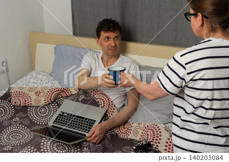 A woman holds out a blue cup to a man who is sitting in bed. He has a laptop in his lap and looks at her. They are in a room at home during the day A woman holds out a blue cup to a man who is sitting in bed. He has a laptop in his lap and looks at her. They are in a room at home during the day 140203084