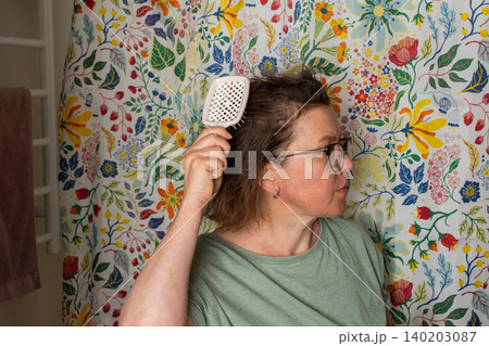 A 50-year-old woman holds a hairbrush while looking in the mirror during her morning bathroom routine. She checks her hair near a floral shower curtain A 50-year-old woman holds a hairbrush while looking in the mirror during her morning bathroom routine. She checks her hair near a floral shower curtain 140203087