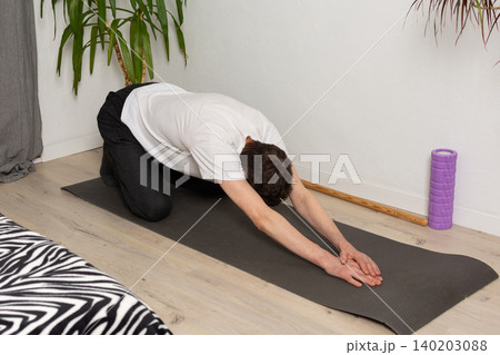 A 53-year-old man performs a stretch on a yoga mat in his home. He reaches forward with his arms extended while sitting on the floor. The room has plants in the background 140203088
