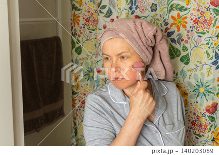 A 50 year old woman applies a facial roller to her face while standing in a bright bathroom. She has a towel wrapped around her hair and wears comfortable clothes A 50 year old woman applies a facial roller to her face while standing in a bright bathroom. She has a towel wrapped around her hair and wears comfortable clothes 140203089