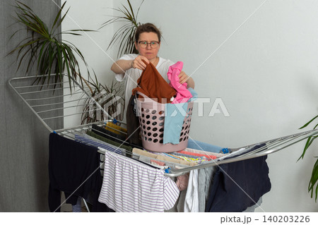 A 52-year-old woman takes freshly cleaned clothes from a drying rack at home. She is focused on her task and handling a basket while surrounded by clothes A 52-year-old woman takes freshly cleaned clothes from a drying rack at home. She is focused on her task and handling a basket while surrounded by clothes 140203226