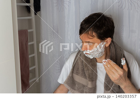 A 53-year-old man is in the bathroom applying shaving cream to his face while preparing for the day. He wears a towel around his neck and focuses on his grooming routine A 53-year-old man is in the bathroom applying shaving cream to his face while preparing for the day. He wears a towel around his neck and focuses on his grooming routine 140203357