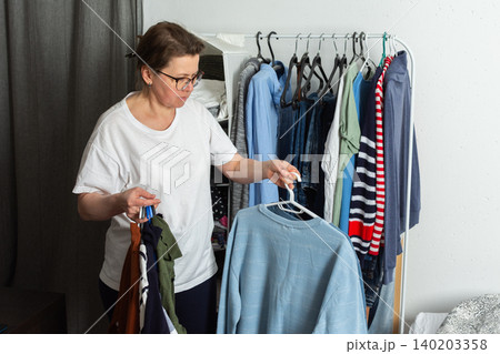Woman sorts and hangs washed clothes on a hanger. She holds a blue sweatshirt and several other items in her hands. This scene takes place in a home environment 140203358