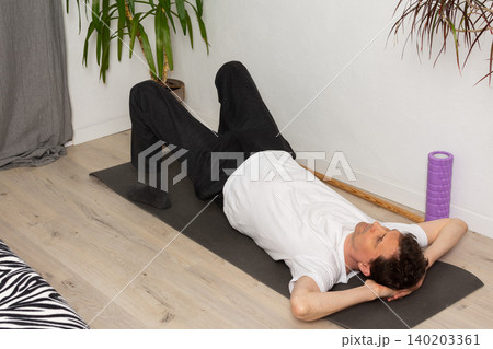 A 53-year-old man performs a leg stretch at home on a mat. He lies on his back with one leg raised and the other leg flat on the ground. He is focused on his workout routine A 53-year-old man performs a leg stretch at home on a mat. He lies on his back with one leg raised and the other leg flat on the ground. He is focused on his workout routine 140203361