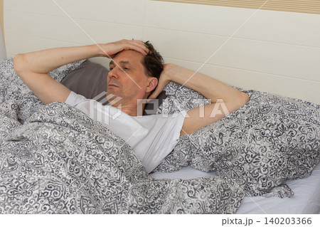A 53-year-old man wakes up in his bedroom and sits up in bed. He holds his head with one hand as he looks thoughtful A 53-year-old man wakes up in his bedroom and sits up in bed. He holds his head with one hand as he looks thoughtful 140203366