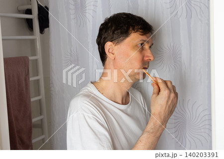 A 53-year-old man stands in his bathroom brushing his teeth. He wears a plain shirt and focuses on his grooming. Morning light filters through the space, creating a daily routine moment A 53-year-old man stands in his bathroom brushing his teeth. He wears a plain shirt and focuses on his grooming. Morning light filters through the space, creating a daily routine moment 140203391