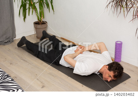 A man, 53 years old, lies on a yoga mat at home, performing a stretching exercise. He focuses on his fitness routine in a simple indoor space with plants around A man, 53 years old, lies on a yoga mat at home, performing a stretching exercise. He focuses on his fitness routine in a simple indoor space with plants around 140203402