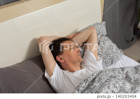 A 53-year-old man is asleep in his bedroom. He is lying on a bed covered with patterned bedding. His arm is stretched above his head. The room is quiet and inviting 140203404
