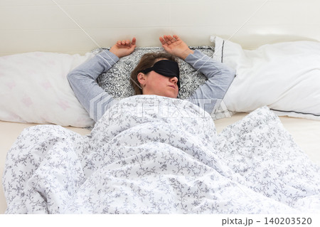A 50-year-old woman is comfortably sleeping in her bed with a sleep mask on. She is wrapped in a soft blanket, resting peacefully in a serene bedroom setting A 50-year-old woman is comfortably sleeping in her bed with a sleep mask on. She is wrapped in a soft blanket, resting peacefully in a serene bedroom setting 140203520