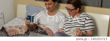 A man and a woman sit together on a bed. They both hold drinks. The man looks at a laptop, while the woman appears to read something on her phone in their home, banner 140203723