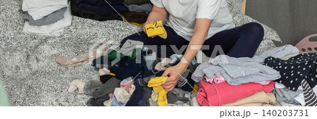A 52-year-old woman sits on her bed with laundry scattered around her. She focuses on sorting socks and organizing other clothes, performing a daily task at home, banner 140203731