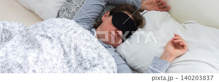A middle-aged woman is sleeping soundly in her bed, wearing a sleep mask. She is surrounded by soft pillows and a warm blanket, taking a break from her daily routine, banner A middle-aged woman is sleeping soundly in her bed, wearing a sleep mask. She is surrounded by soft pillows and a warm blanket, taking a break from her daily routine, banner 140203837