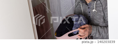 A 53-year-old man removes clean clothes from the washing machine and places them into a basket. He stands in a bathroom with a simple design and light colors, banner A 53-year-old man removes clean clothes from the washing machine and places them into a basket. He stands in a bathroom with a simple design and light colors, banner 140203839