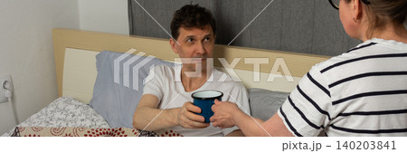 A woman holds out a blue cup to a man who is sitting in bed. He has a laptop in his lap and looks at her. They are in a room at home during the day, banner A woman holds out a blue cup to a man who is sitting in bed. He has a laptop in his lap and looks at her. They are in a room at home during the day, banner 140203841