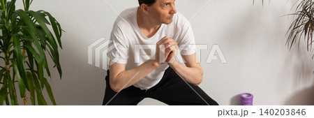 A 53-year-old man practices exercises at home on a yoga mat. He focuses on his strength and flexibility while avoiding distractions from the surroundings. Plants stand around him, banner A 53-year-old man practices exercises at home on a yoga mat. He focuses on his strength and flexibility while avoiding distractions from the surroundings. Plants stand around him, banner 140203846
