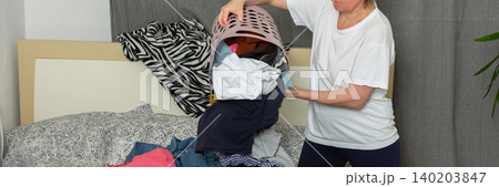 A 52-year-old woman pours laundry out of a basket onto her bed. She sorts through the clothes in her comfortable room as part of her routine tasks, banner A 52-year-old woman pours laundry out of a basket onto her bed. She sorts through the clothes in her comfortable room as part of her routine tasks, banner 140203847