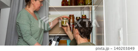 Married couple places jars of canned goods onto shelves in their pantry. They are inside their home with a view of flowers and the city from the balcony. It is autumn, banner 140203849