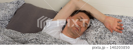A 53-year-old man is asleep in his bedroom. He is lying on a bed covered with patterned bedding. His arm is stretched above his head. The room is quiet and inviting, banner 140203850