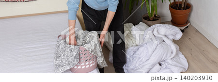 A 53-year-old man is in his bedroom folding and changing bed linen. He is holding a laundry basket while arranging the sheets on the bed. Plants are visible in the room, banner A 53-year-old man is in his bedroom folding and changing bed linen. He is holding a laundry basket while arranging the sheets on the bed. Plants are visible in the room, banner 140203910