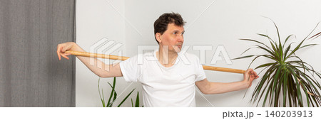 A 53-year-old man is working out at home. He is using a stick for balance and is focused on his exercise. The room features plants and a yoga mat on the floor, banner A 53-year-old man is working out at home. He is using a stick for balance and is focused on his exercise. The room features plants and a yoga mat on the floor, banner 140203913