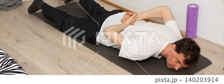 A man, 53 years old, lies on a yoga mat at home, performing a stretching exercise. He focuses on his fitness routine in a simple indoor space with plants around, banner A man, 53 years old, lies on a yoga mat at home, performing a stretching exercise. He focuses on his fitness routine in a simple indoor space with plants around, banner 140203914