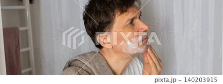 A 53-year-old man stands in the bathroom applying skincare on his face. He wears a towel around his neck and is focused on his morning routine. The time is early morning, banner A 53-year-old man stands in the bathroom applying skincare on his face. He wears a towel around his neck and is focused on his morning routine. The time is early morning, banner 140203915